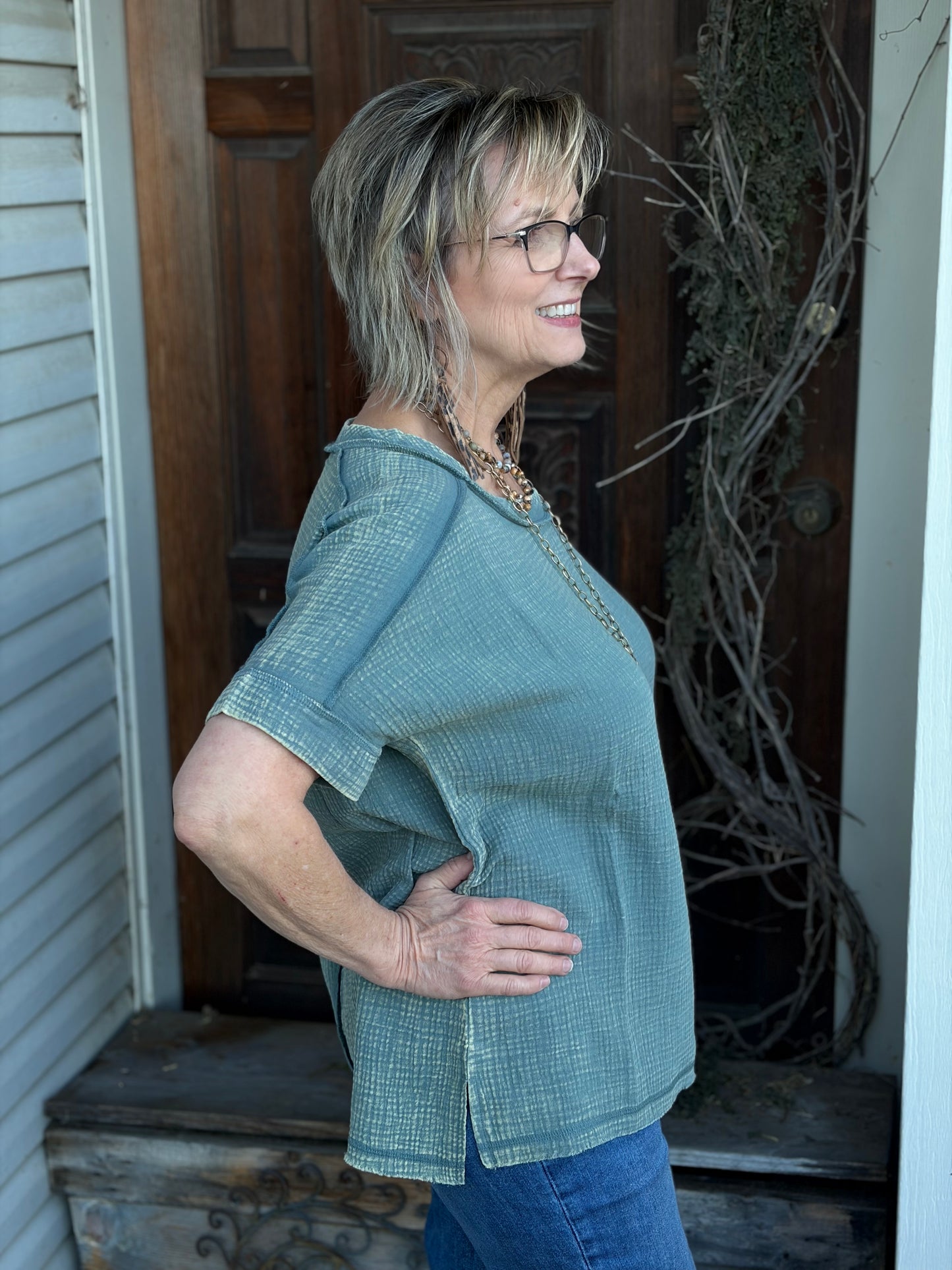 Woman wearing a sage top and blue jeans standing in front of a wooden door.