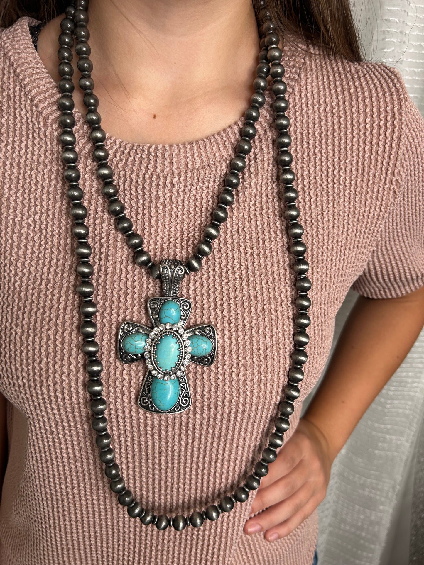 Necklace with a cross pendant featuring turquoise stones on a person wearing a pink sweater.