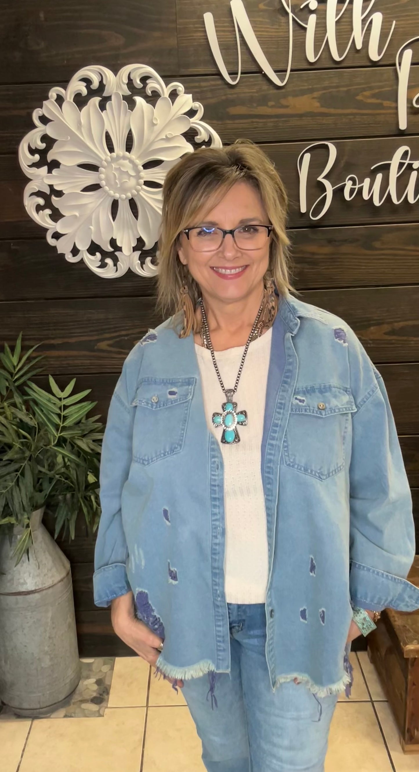 Woman wearing a denim jacket with a cross necklace in front of a decorative wall.