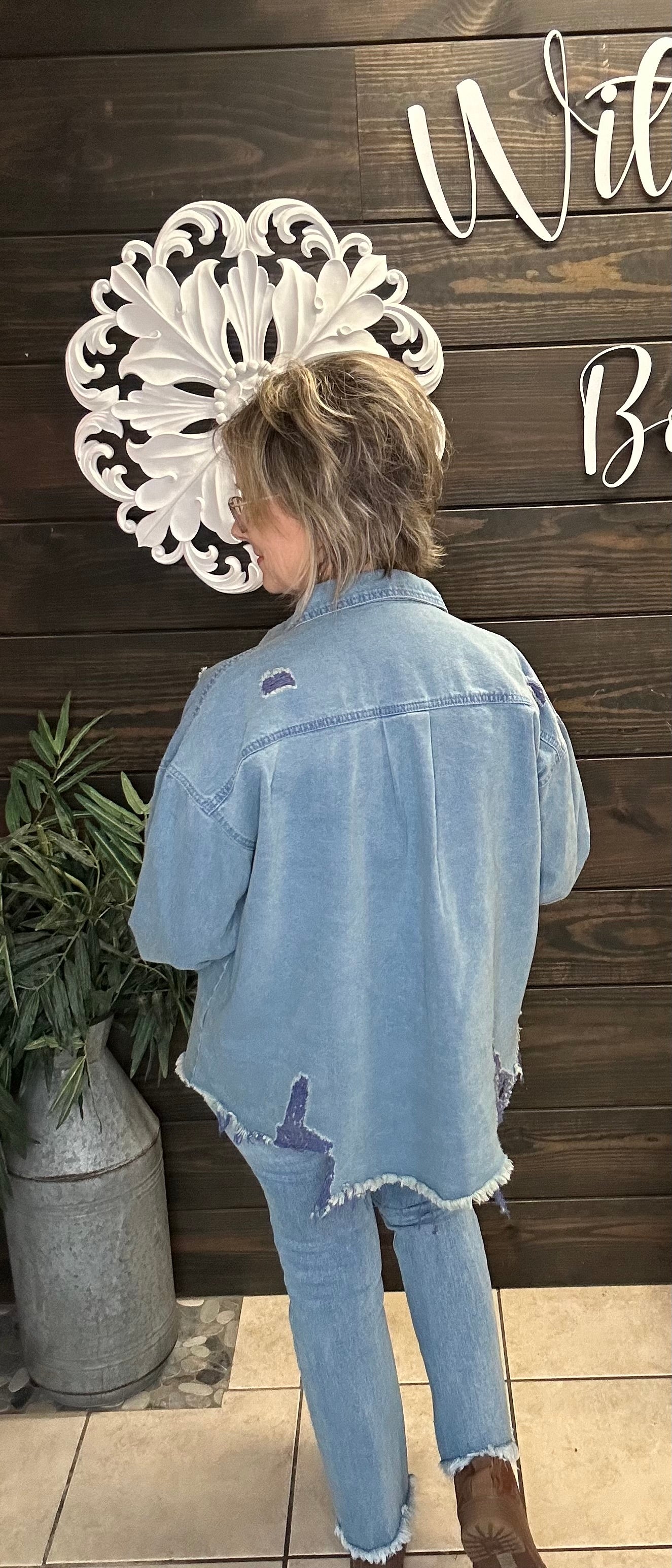 Person wearing a light blue denim jacket and jeans standing in front of a wooden wall with decorative elements.