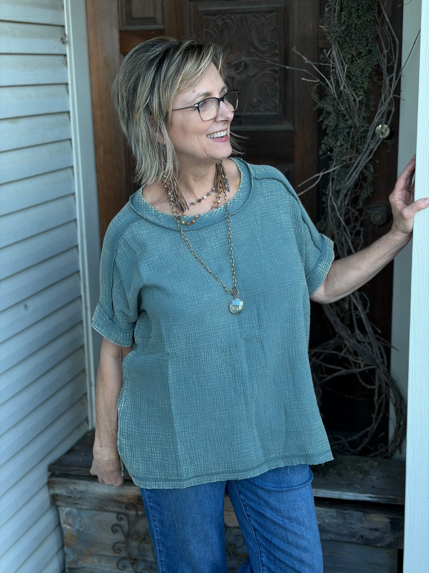 Woman wearing a sage shirt and blue jeans standing on a porch.