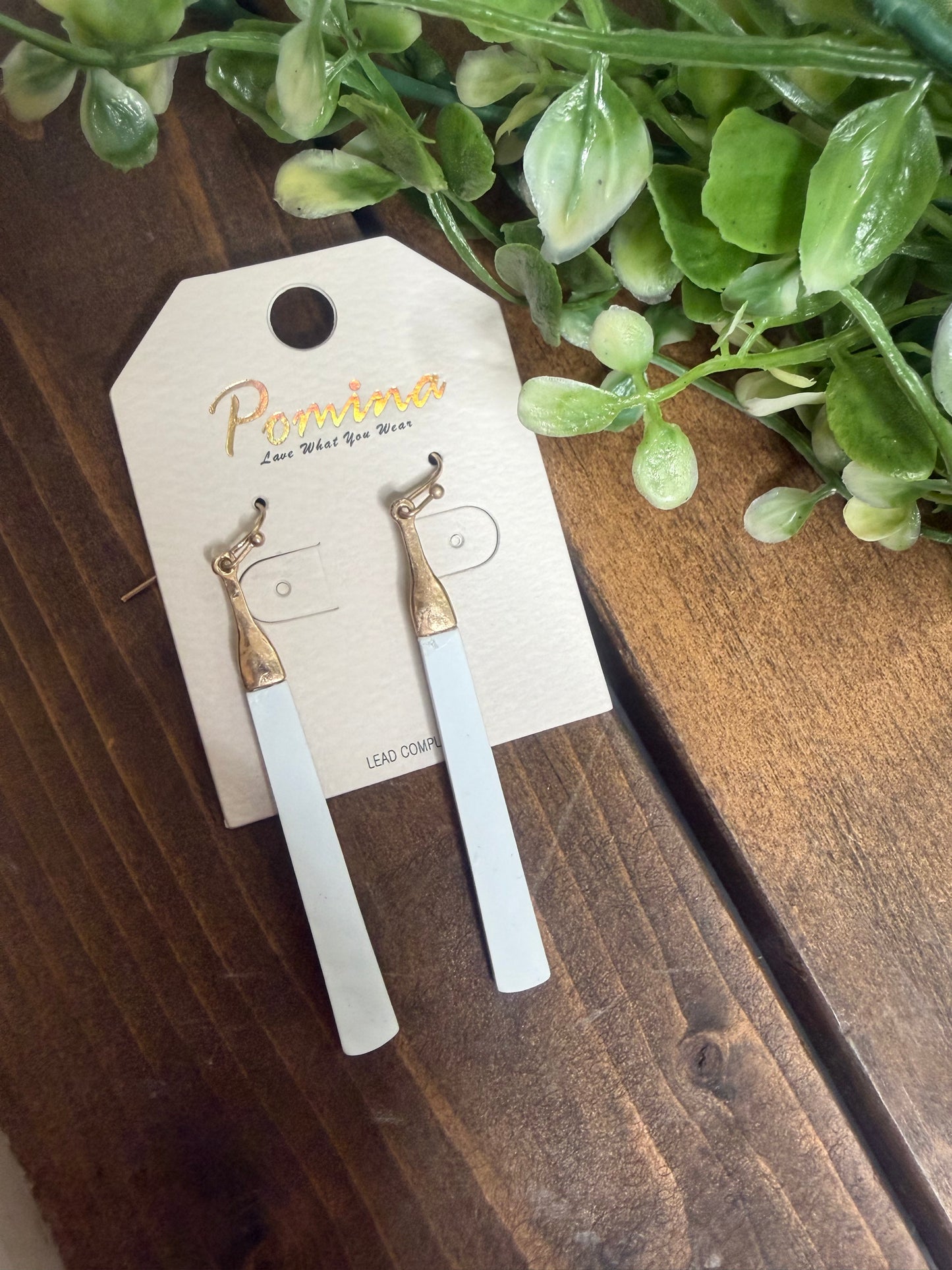 A pair of long white earrings with gold accents at the top on wooden background with greenery