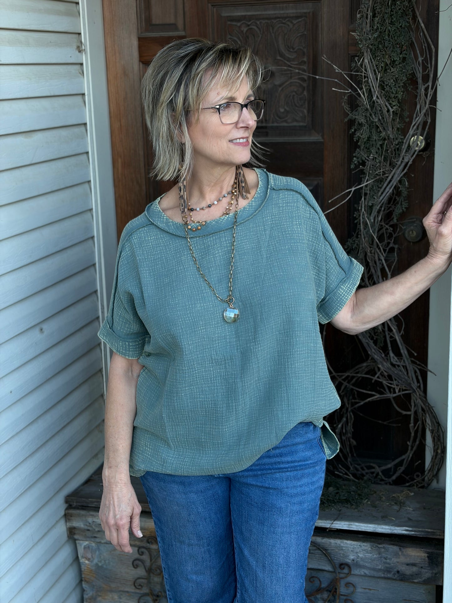 Woman wearing a green tunic and blue jeans standing on a porch.