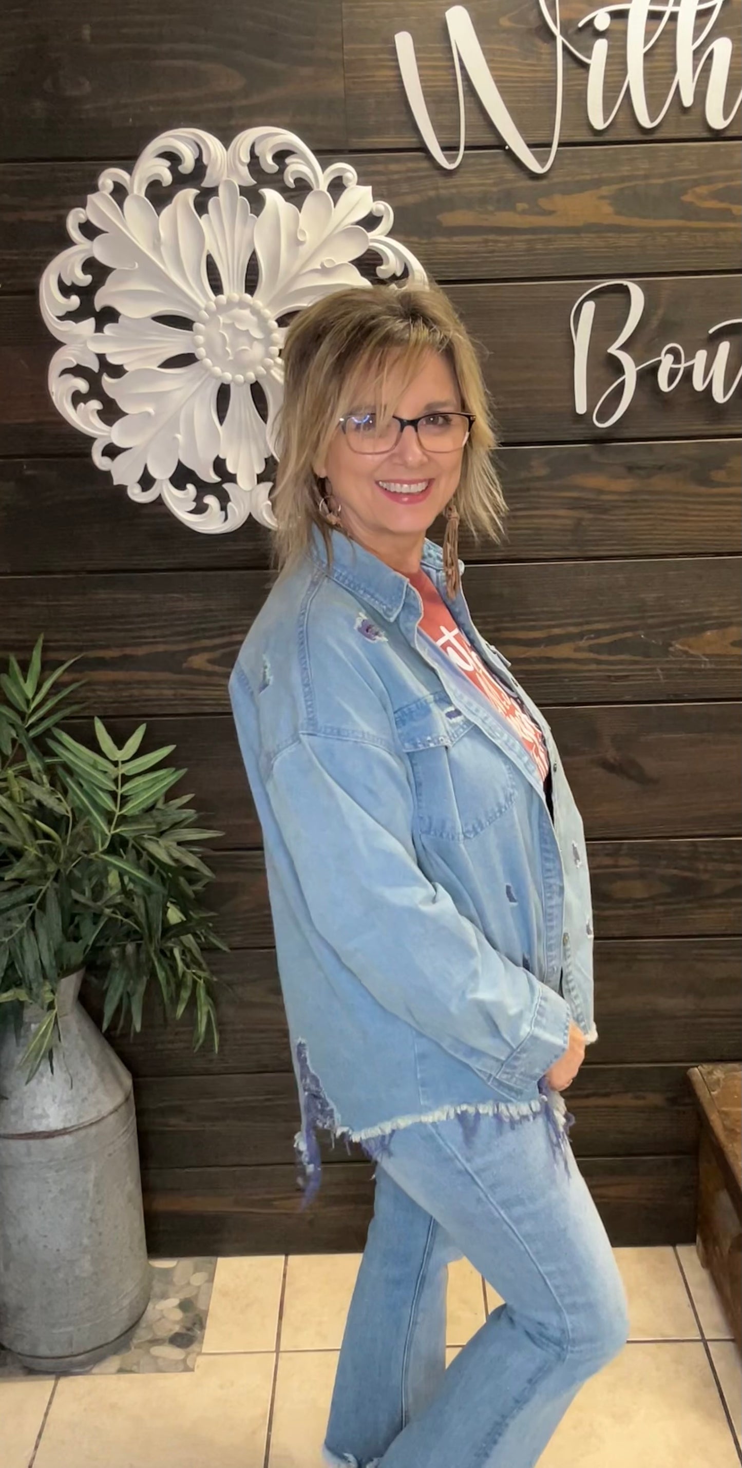 Woman wearing a denim jacket and jeans standing in front of a decorative wall with text.