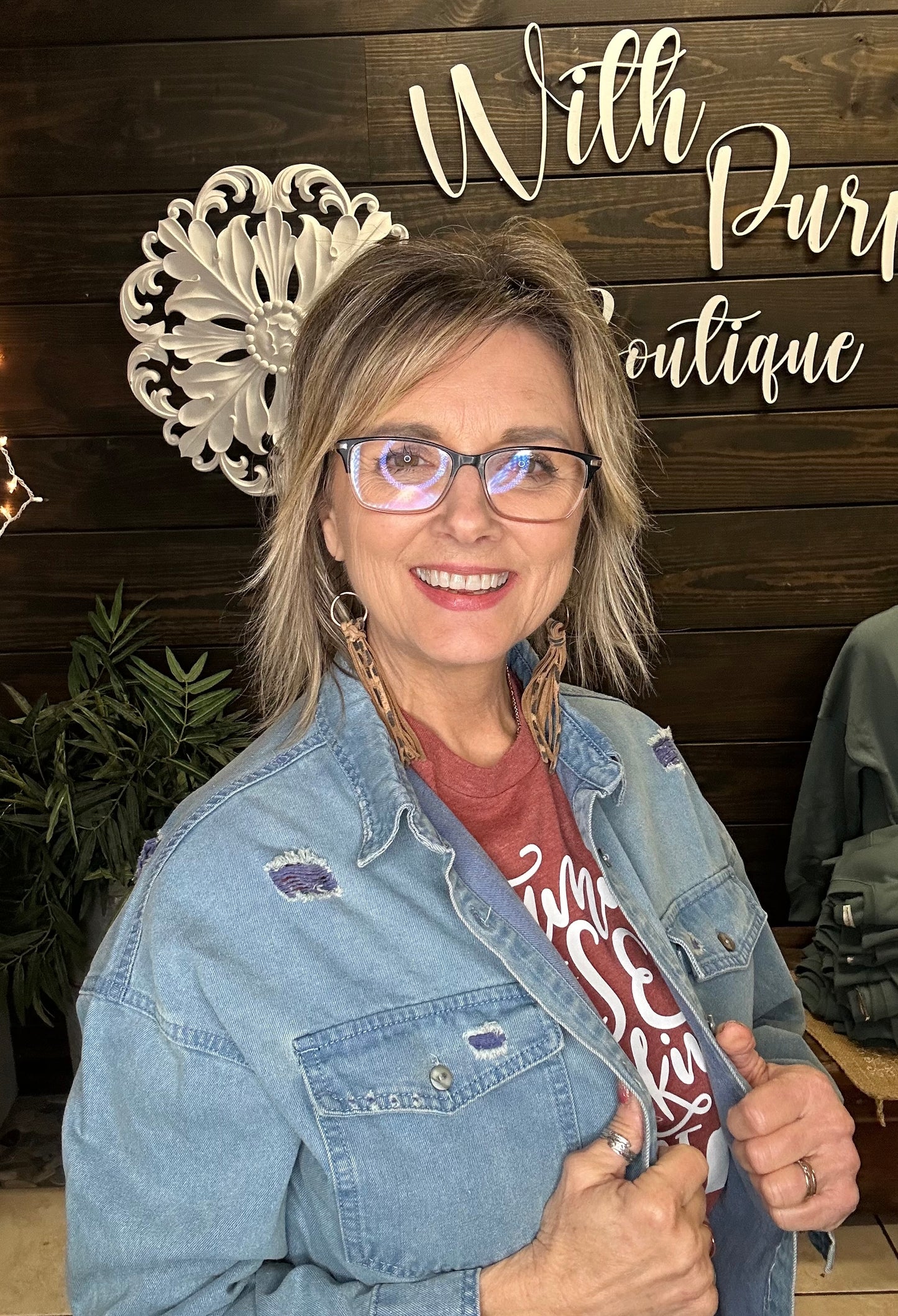 Woman wearing glasses and a denim jacket in front of a wooden wall with decorative elements.