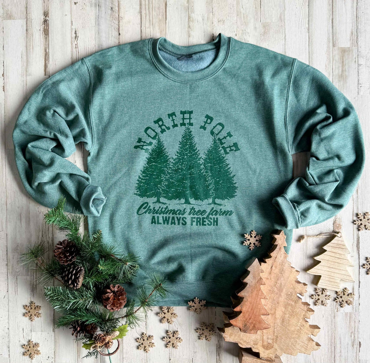 Green sweatshirt with Christmas tree design and text on a wooden surface with decorative elements.