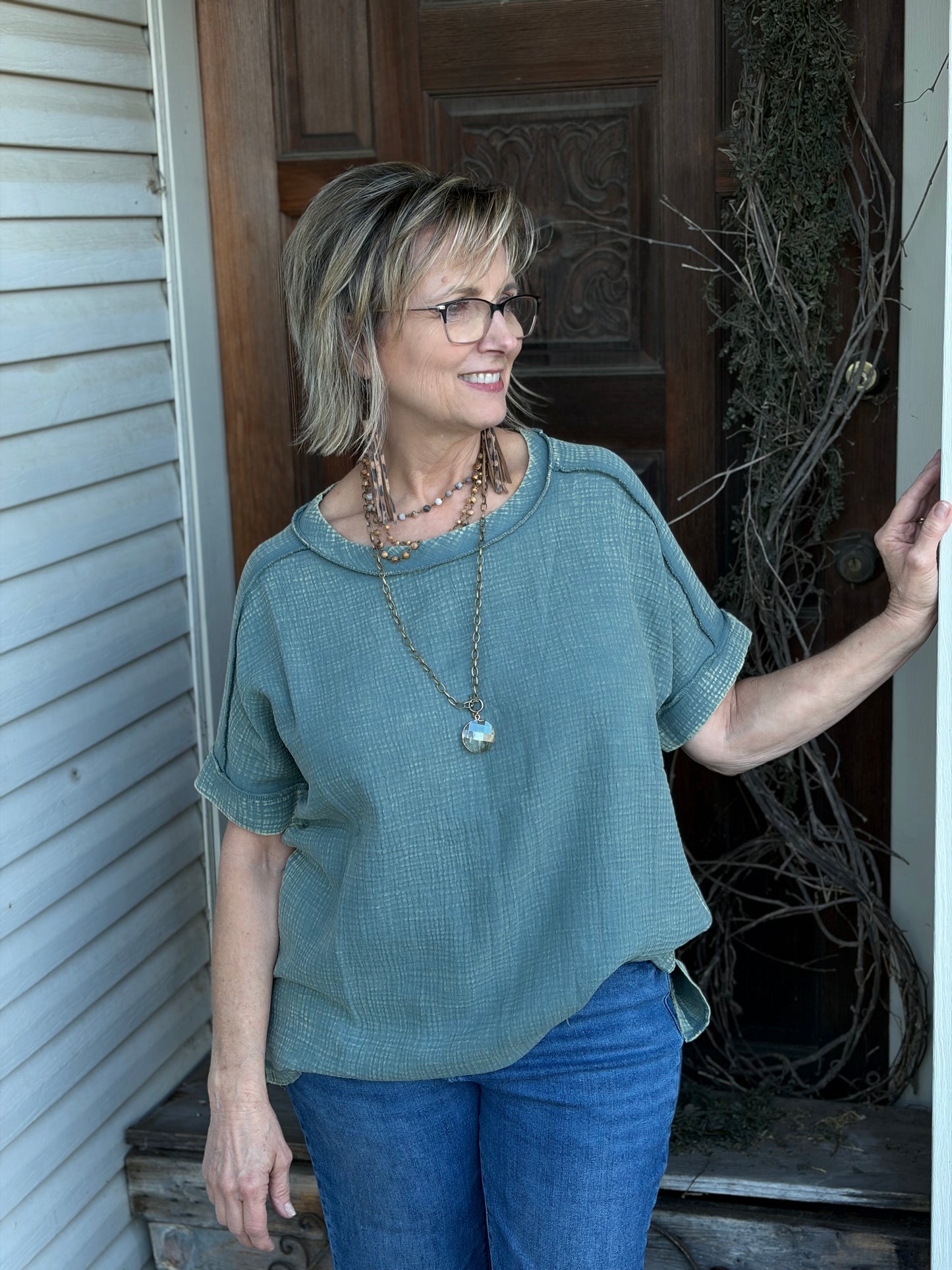 Woman wearing a sage tunic and blue jeans standing outside a house.