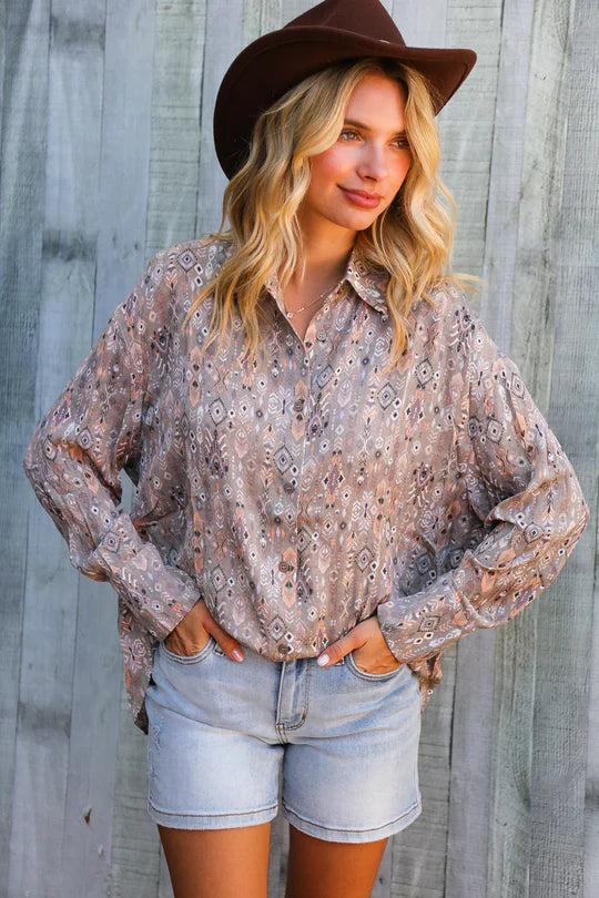 Woman wearing a patterned shirt, denim shorts, and a brown hat against a wooden background