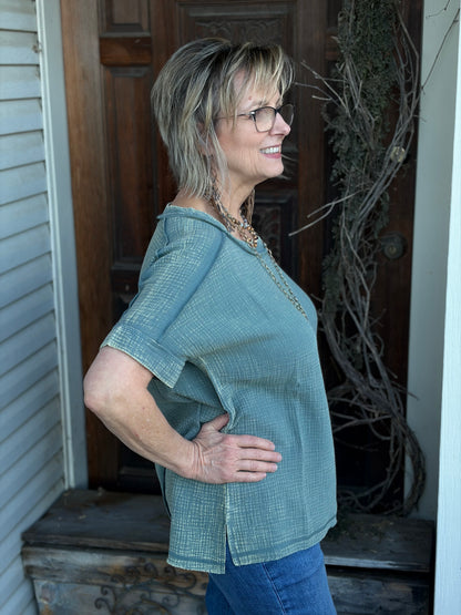 Woman wearing a sage top and blue jeans standing in front of a wooden door.