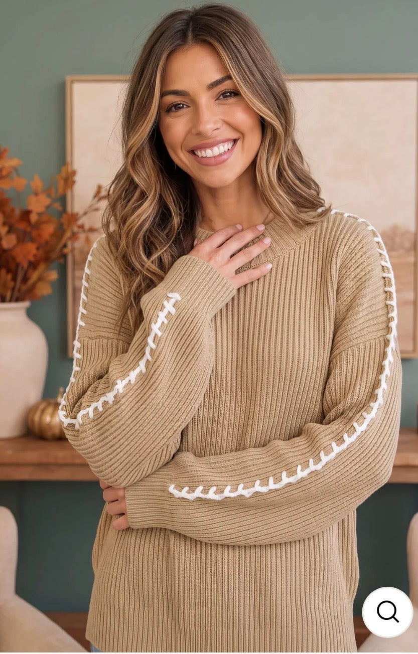 A woman wearing a beige ribbed knit sweater with white decorative  whipstitching along the sleeve, posing in a cozy indoor setting.

