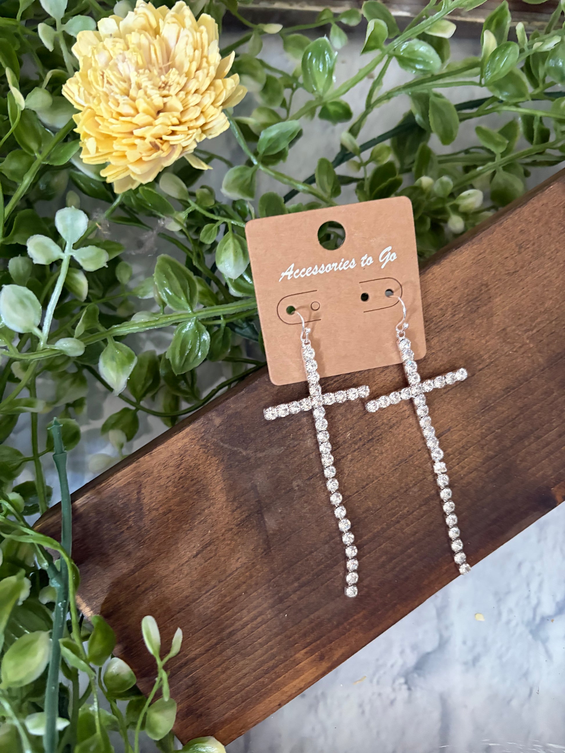 Cross-shaped dangle earrings with clear rhinestones on a tan display card, styled on a wooden board with green leaves and a yellow flower.
