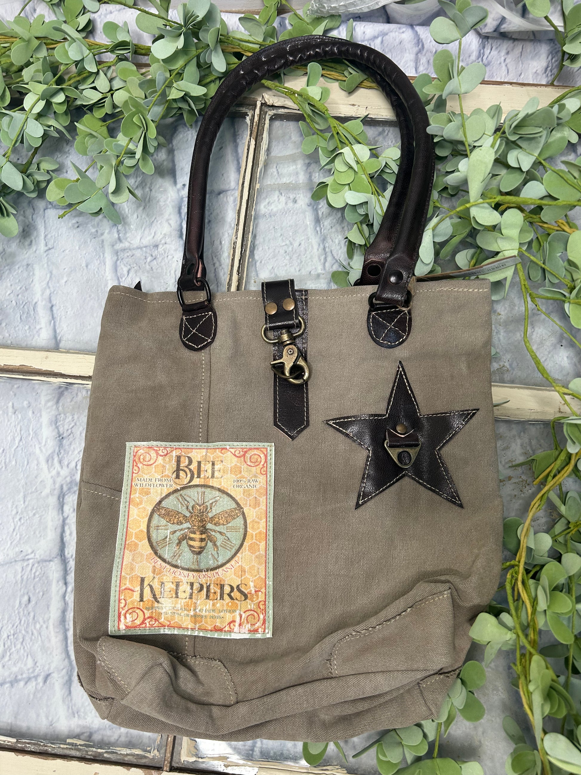 This uniquely charming tote is crafted from upcycled canvas for a vintage vibe. Attention to detail shows in the quality, including lobster claw clips and a back zippered pocket, three inside pockets, star applique and bee patch inlay!&nbsp; (Bee-cause who doesn't want a bag with buzz?)&nbsp;

Designed &amp; Manufactured in USA/Hand Crafted bags made from upcycled canvas 
such as repurposed military tents &amp; more /approx:10 x 14 x 3.5