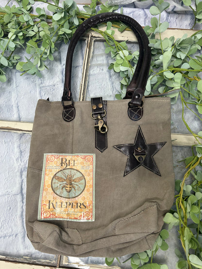 This uniquely charming tote is crafted from upcycled canvas for a vintage vibe. Attention to detail shows in the quality, including lobster claw clips and a back zippered pocket, three inside pockets, star applique and bee patch inlay!&nbsp; (Bee-cause who doesn't want a bag with buzz?)&nbsp;

Designed &amp; Manufactured in USA/Hand Crafted bags made from upcycled canvas 
such as repurposed military tents &amp; more /approx:10 x 14 x 3.5