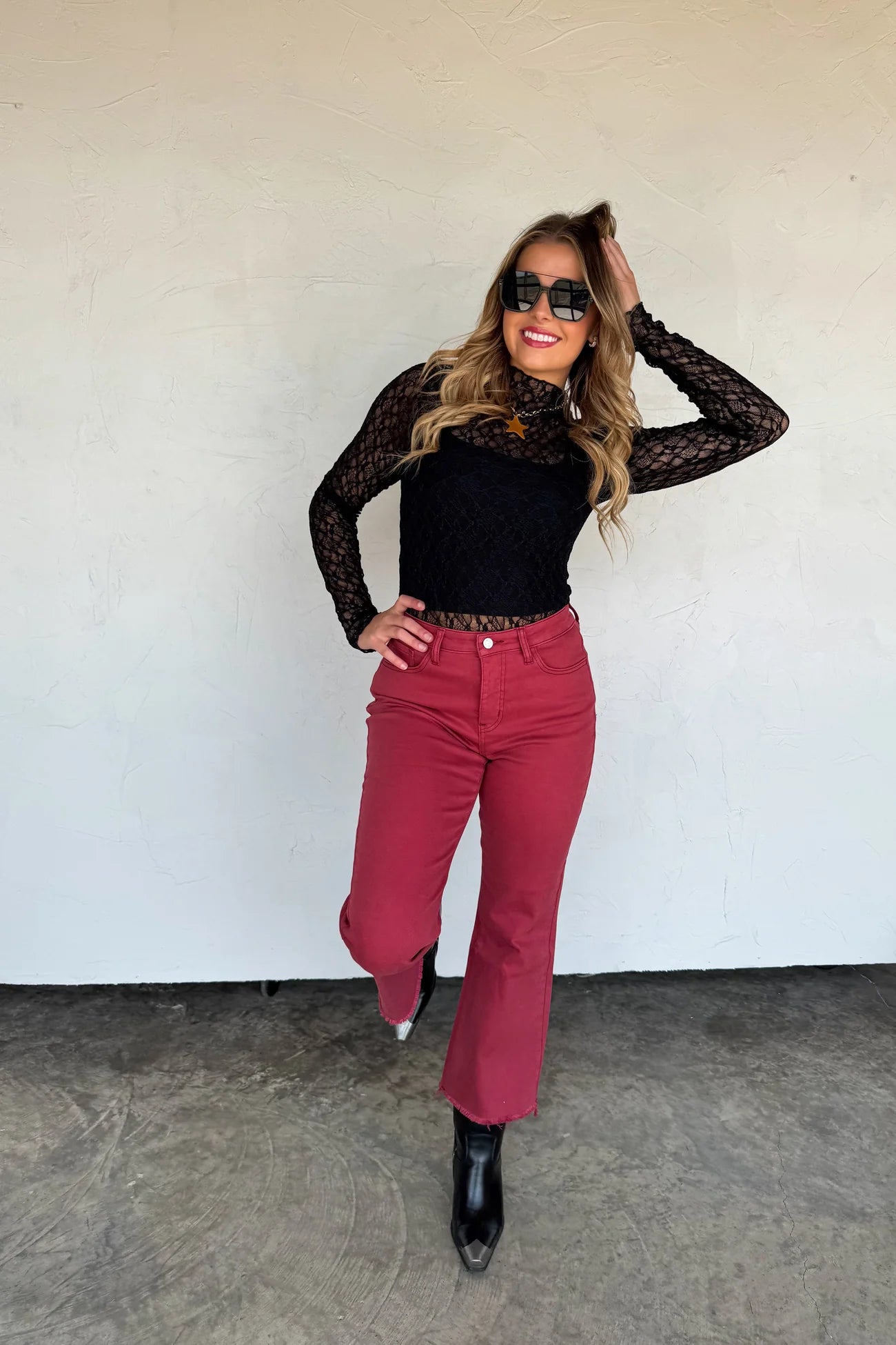 Woman wearing a black lace top and maroon pants against a white wall.