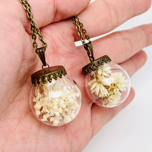 Two glass pendant necklaces with dried flowers inside, held in a hand.