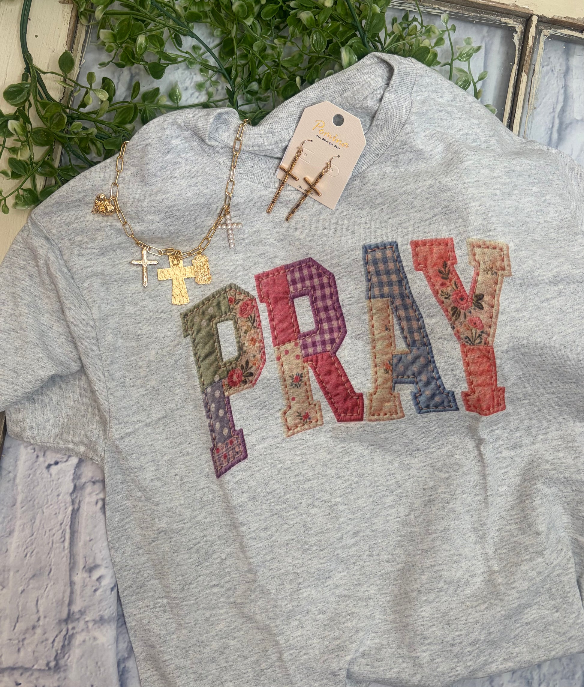 Gray tee shirt with 'PRAY' in colorful quilted looking letters and gold necklace on a textured surface.