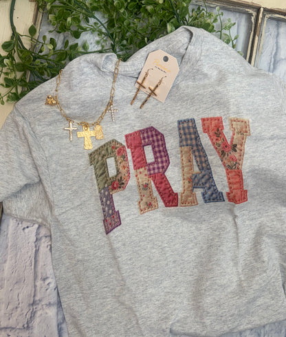 Gray tee shirt with 'PRAY' in colorful quilted looking letters and gold necklace on a textured surface.