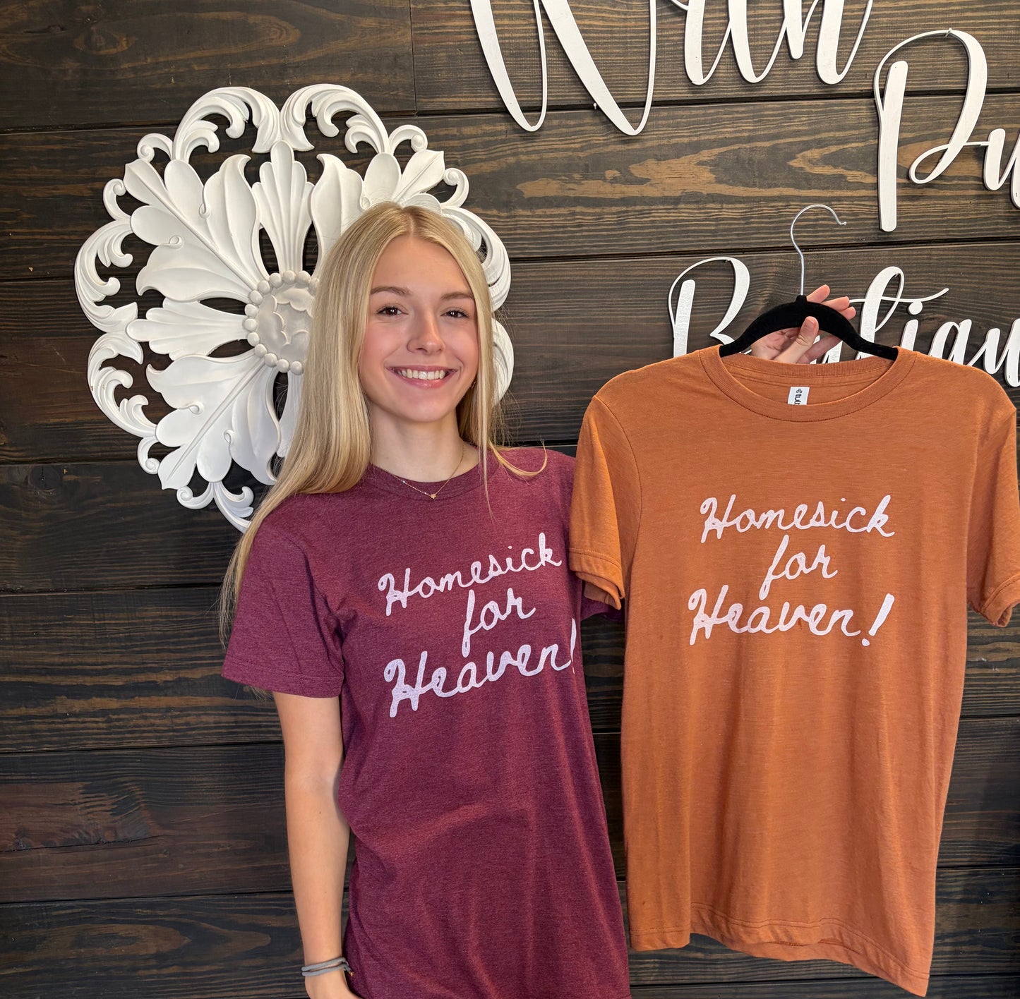 Woman wearing a maroon t-shirt with 'Homesick for Heaven' text, next to an orange t-shirt with the same text on a wooden background.