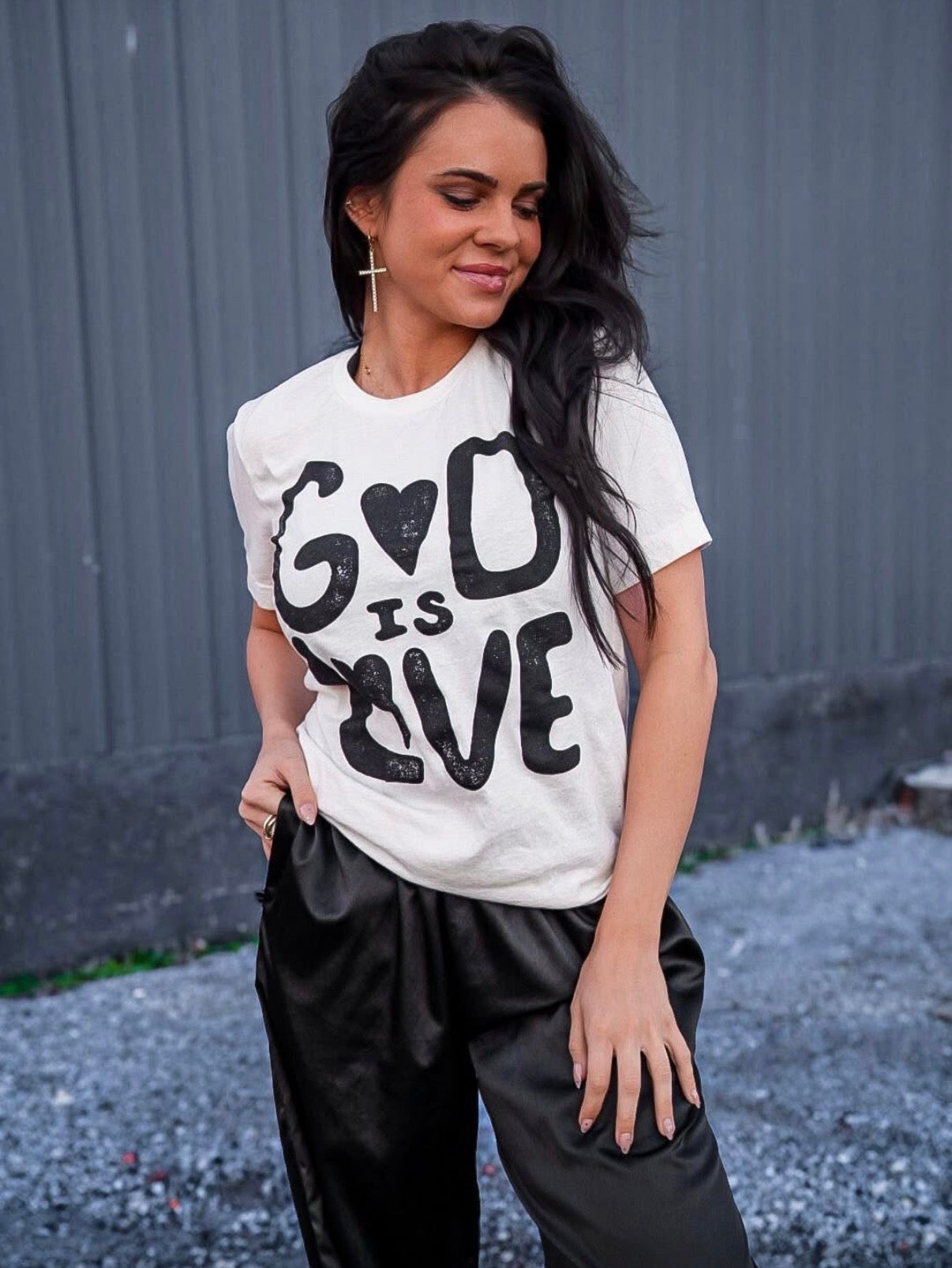 Woman wearing a white t-shirt with 'God is Love' text against a gray wall.