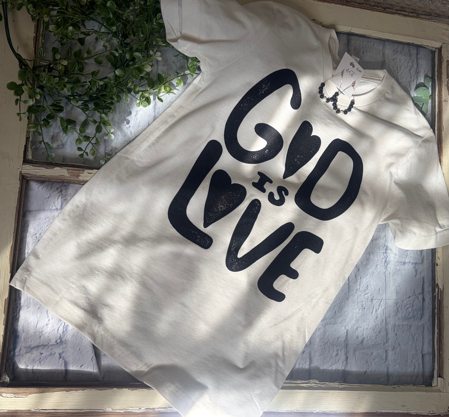 White t-shirt with 'God is Love' text on a textured surface with plants in the background
