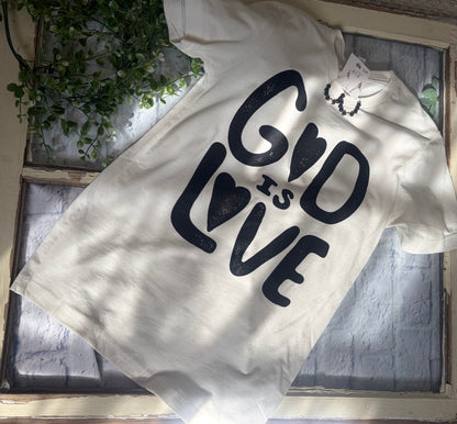 White t-shirt with 'God is Love' text on a textured surface with plants in the background