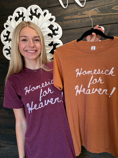 Woman wearing a purple t-shirt with 'Homesick for Heaven' text, next to an orange t-shirt on a hanger.