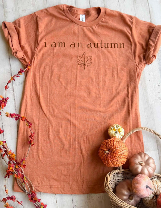 Copper-colored t-shirt with 'I am an autumn' text and leaf design, placed on a wooden surface with pumpkins and leaves.