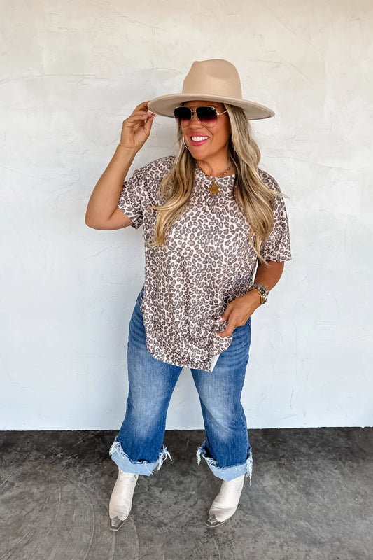 Woman wearing a patterned cheetah top, jeans, white boots, and a beige hat against a plain background