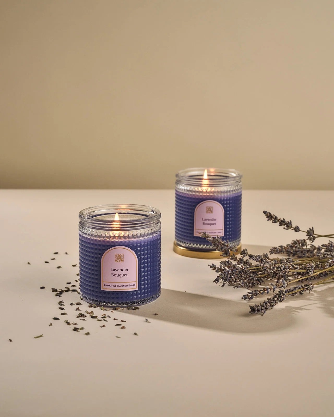Two lavender-scented candles with a label on a beige surface, accompanied by dried lavender flowers.