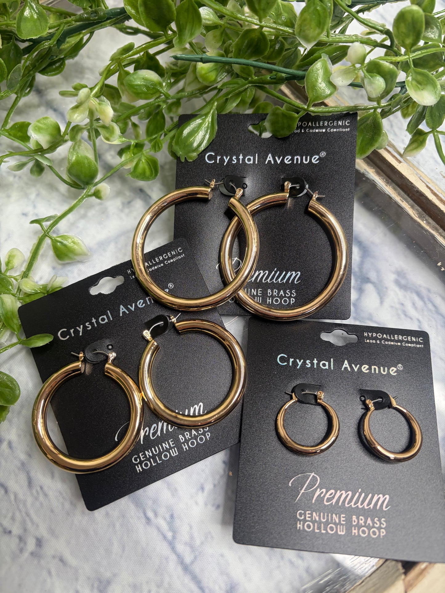 Gold hoop earrings in 3 sizes on black cards with 'Crystal Avenue' branding, surrounded by green leaves.