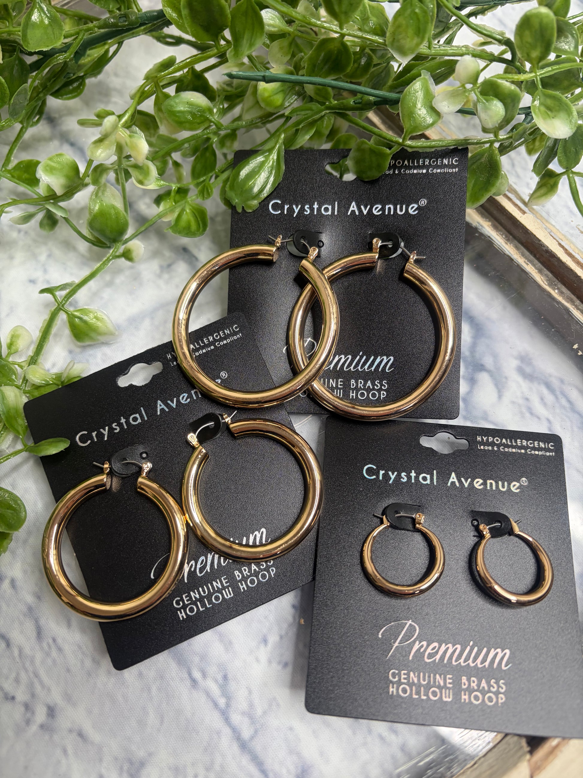 Gold hoop earrings in 3 sizes on black cards with 'Crystal Avenue' branding, surrounded by green leaves.