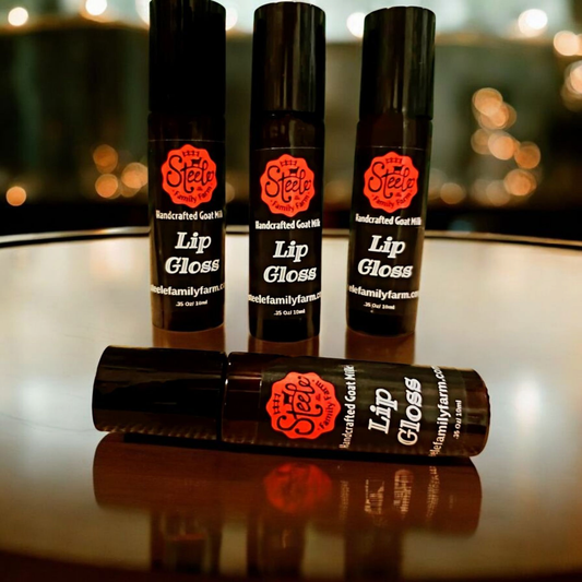 Four bottles of lip gloss from Steel Family Farm on a reflective surface with blurred lights in the background.