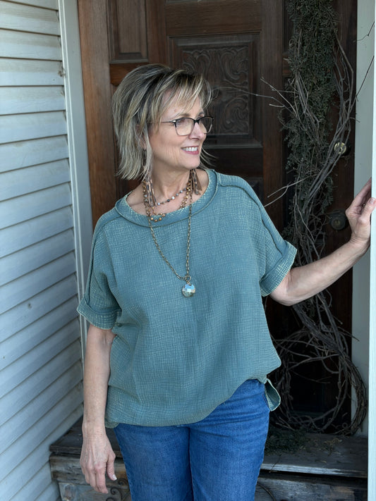 Woman wearing a sage tunic and blue jeans standing outside a house.