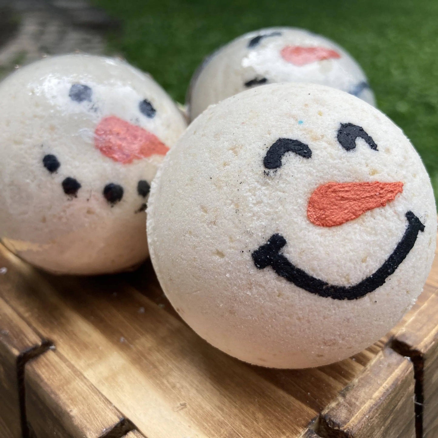 Three snowman-themed bath bombs on a wooden surface with a grassy background.