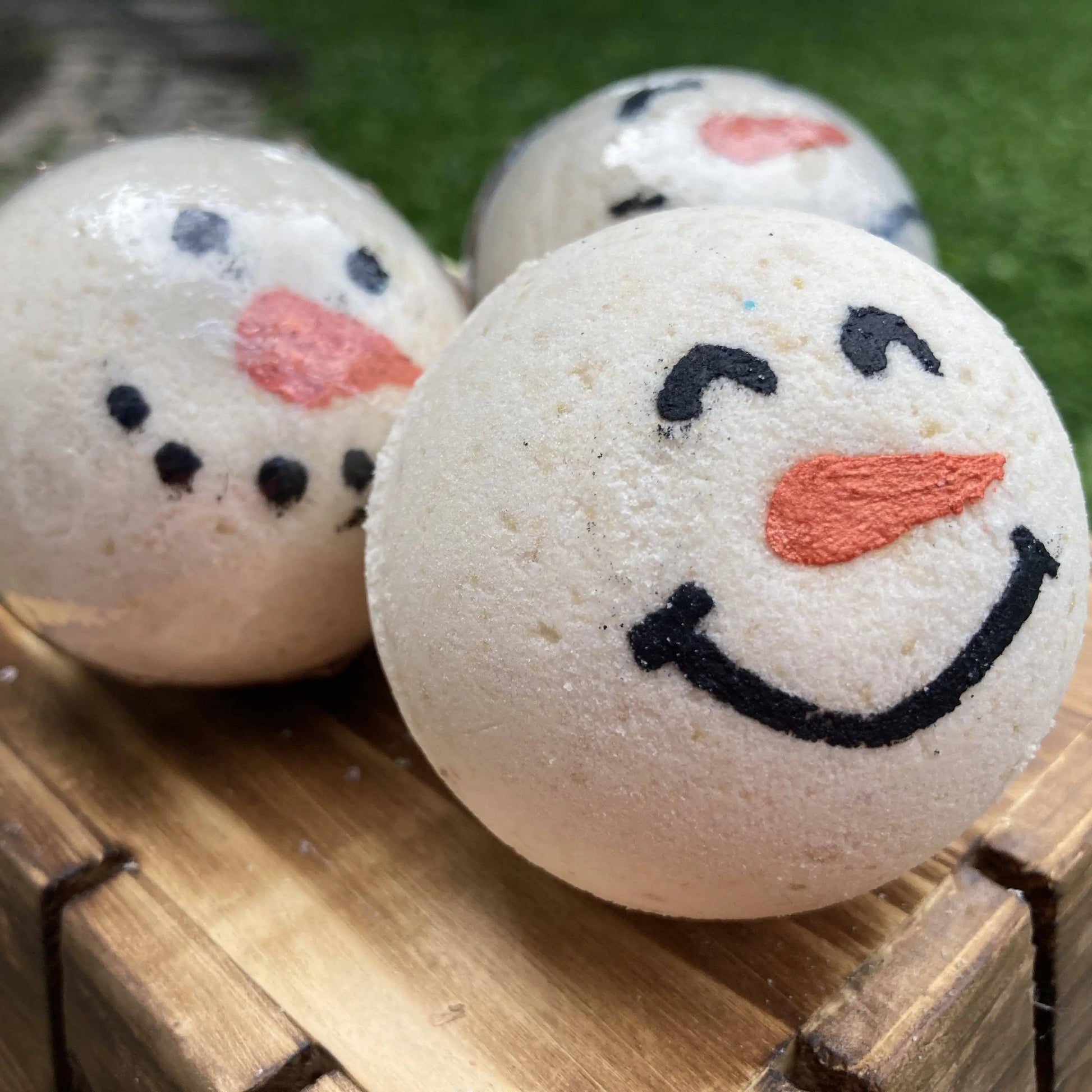 Three snowman-themed bath bombs on a wooden surface with a grassy background.