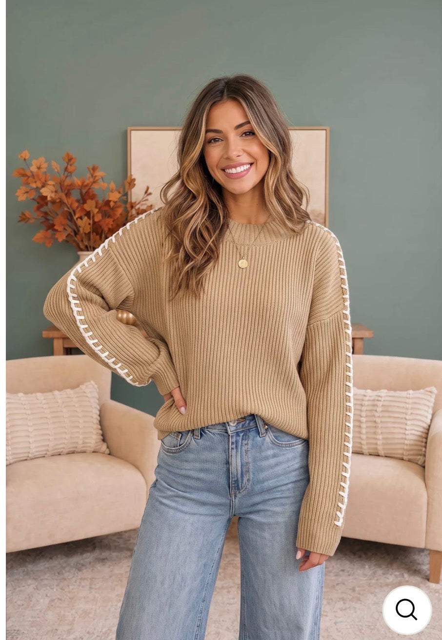 A woman wearing a beige ribbed knit sweater with white lace-up trim on the sleeves, paired with light blue jeans, standing in a cozy living room setting.