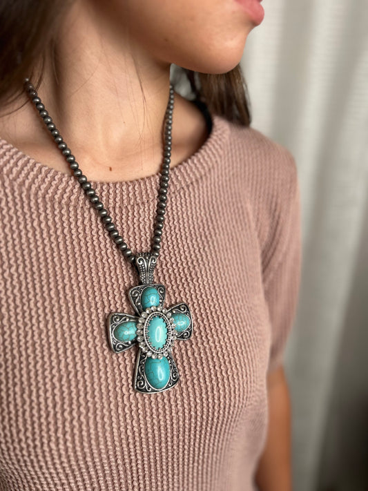 Necklace with a cross pendant featuring turquoise stones worn by a person in a pink sweater.
