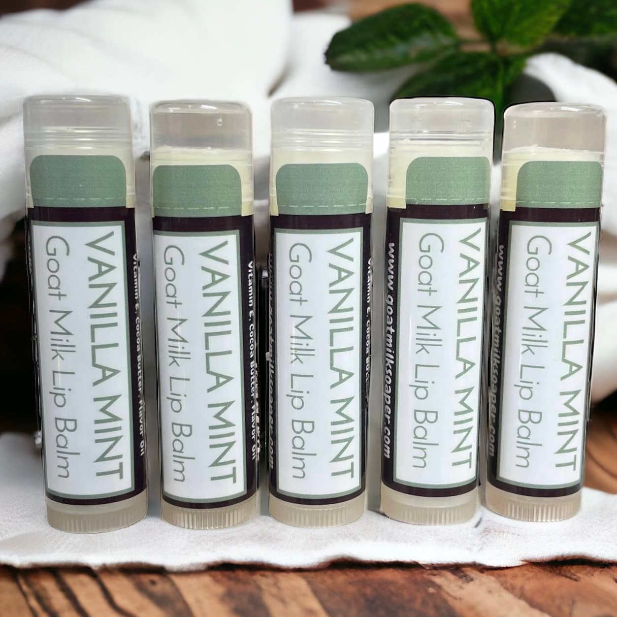 Five tubes of Vanilla Mint Goat Milk Lip Balm on a white cloth with a wooden surface and green leaves in the background.