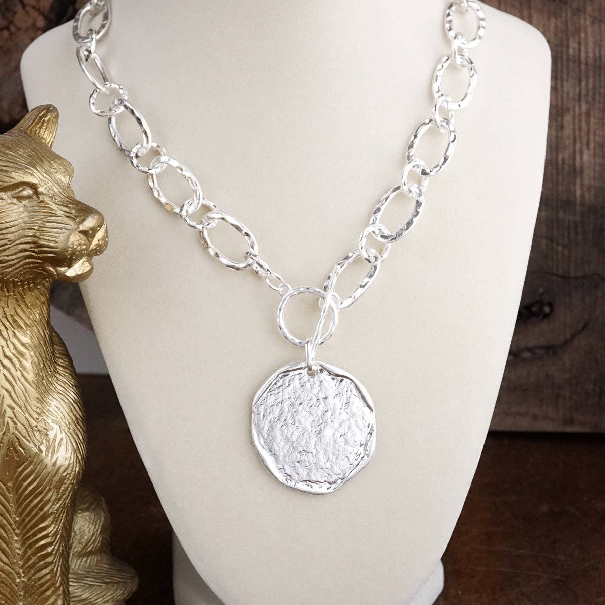 Silver chain necklace with a textured pendant on a mannequin bust, next to a gold animal figurine.