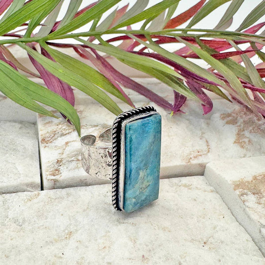 Part of the Kashi Collection, this ring features a prominent, genuine Apatite stone set in silver-plated brass. The Kashi collection highlights the natural beauty of semi-precious stones through traditional craftsmanship. The adjustable band ensures a flexible fit for a wide range of ring sizes. As Apatite is a naturally occurring stone, expect unique variations in its deep teal color and inclusions, ensuring every ring is distinctly its own. Specifications: Material: Apatite, Silver-plated brass Stone Shap