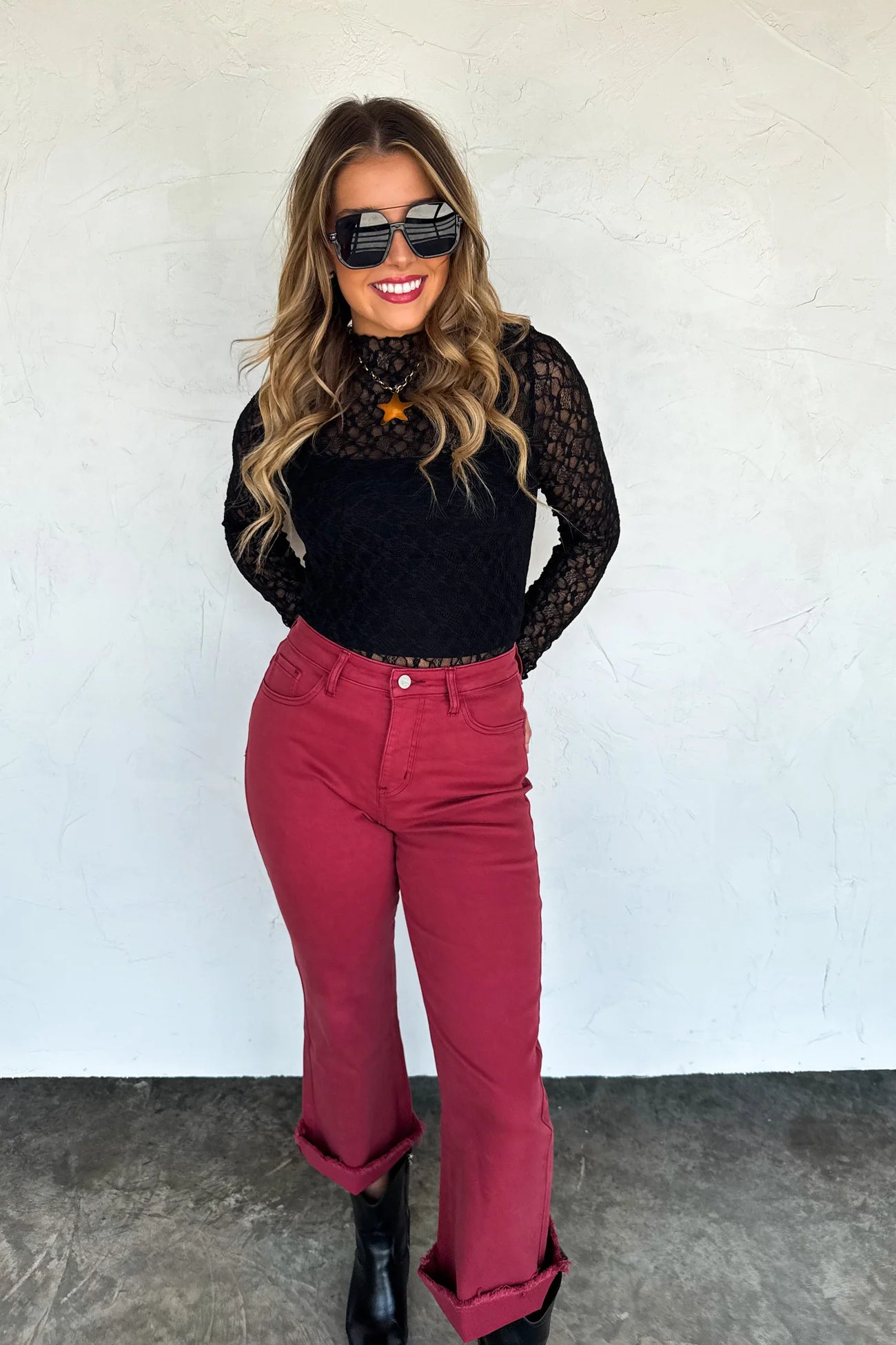 Woman wearing a black top with sheer sleeves and maroon pants against a white wall.